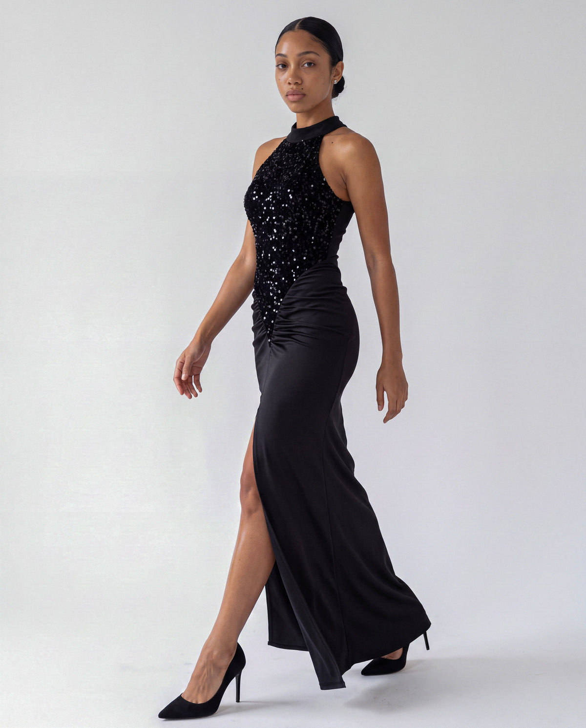 Velvet Sequin High Neck Maxi Dress in black, walking, showing the sequin halter bodice, ruched satin skirt and high front slit