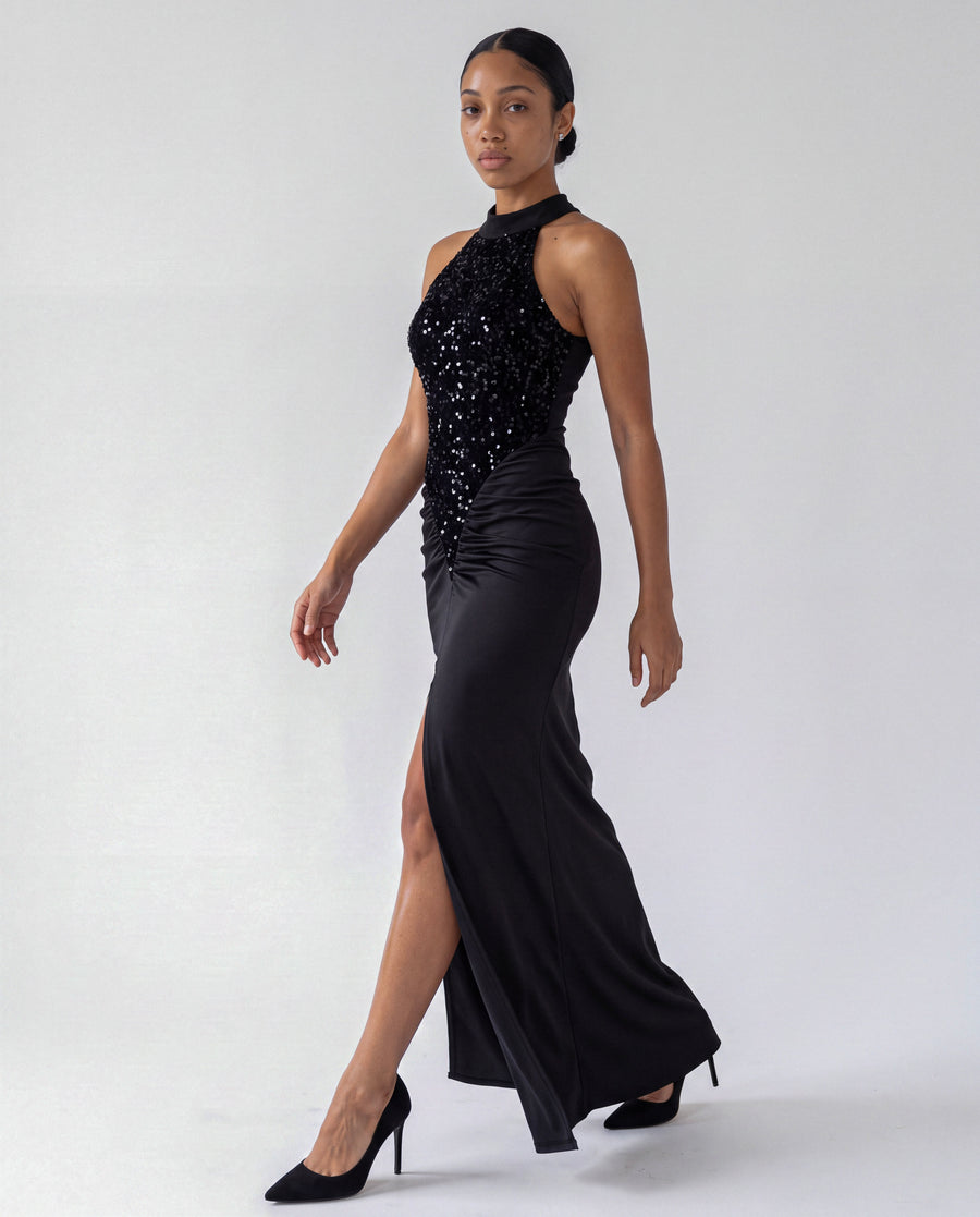 Velvet Sequin High Neck Maxi Dress in black, walking, showing the sequin halter bodice, ruched satin skirt and high front slit